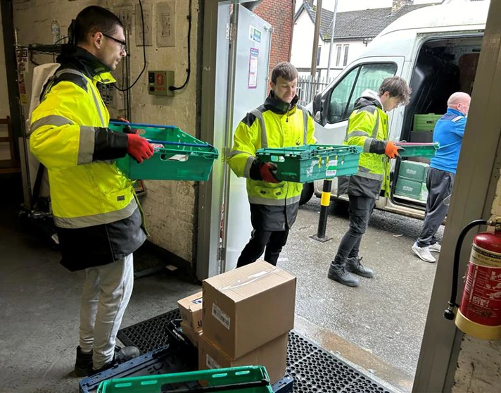 Three young men in high-visibility jackets unloading green crates from a delivery van at a warehouse entrance. Three young men in high-visibility jackets unloading green crates from a delivery van at a warehouse entrance.